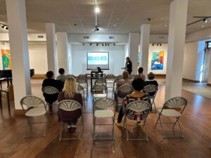 Cheryl Maxsom presenting in front of a screen in an art gallery room to a few rows about 10 people total.